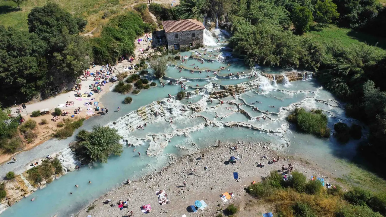 Letecký pohled na termální kaskády Cascate del Mulino v Saturnii, Itálie. Z výšky se odhaluje jedinečná struktura přírodních bazénků s tyrkysovou vodou, obklopených vápencovými terasami a nádhernou toskánskou krajinou. Termální prameny, vyvěrající z hlubin země, vytvářejí fascinující kontrast mezi bílými skalami a sytě modrou vodou. Jedna z nejkrásnějších přírodních scenérií v Itálii.