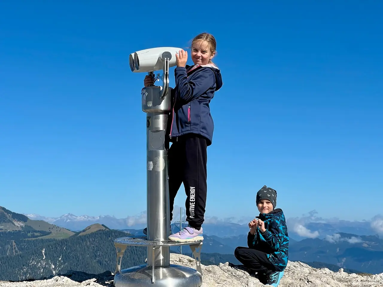 Dcera se dívá vyhlídkovým dalekohledem na Monte Lussari, syn sedí vedle na skále