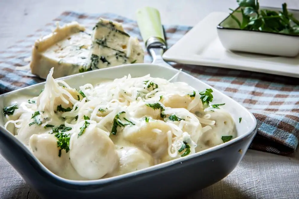 Čerstvě uvařené italské gnocchi se sýrem a bylinkami v bílé misce, vedle modro-bílého sýra s nožem