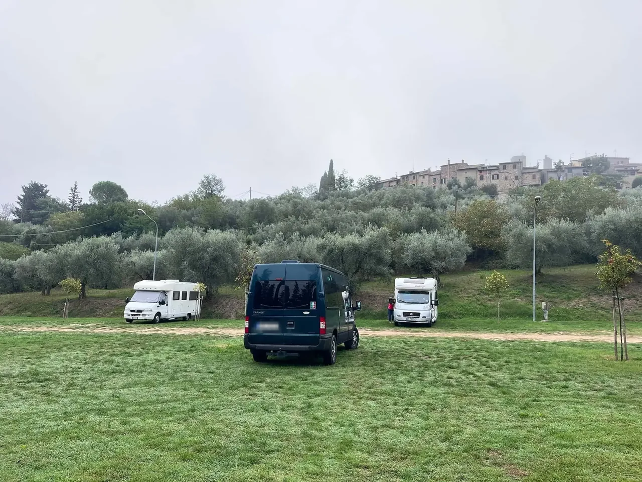 Naše černé rodinné auto zaparkované na travnatém kempingu v Lucignano mezi dalšími obytňáky pod olivovníky