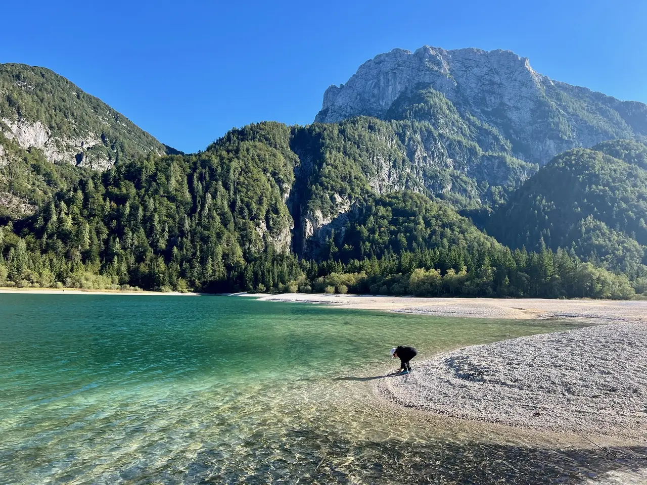 Tyrkysová voda Lago del Predil u oblázkové pláže, nad jezerem zalesněné svahy a skalnaté štíty