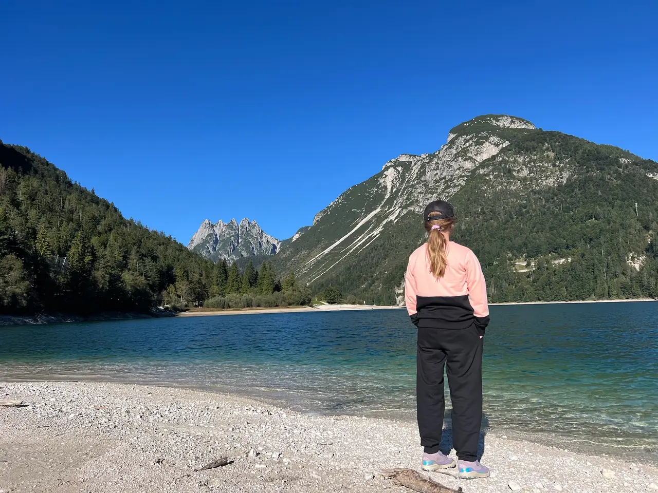 Dcera stojí na oblázkovém břehu Lago del Predil a dívá se na hory nad jezerem