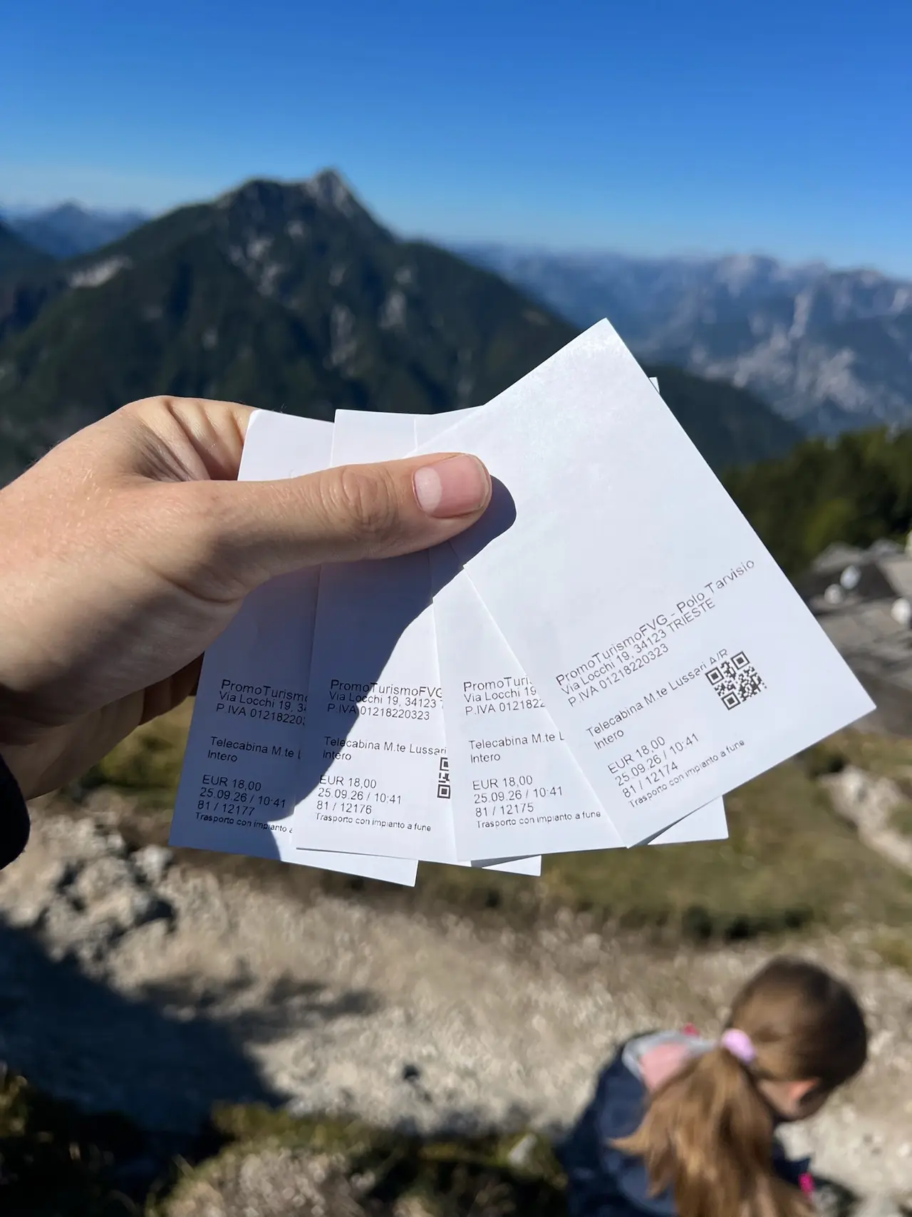 Čtyři zpáteční jízdenky na lanovku Monte Lussari za 18 eur v ruce, v pozadí hory a dcera