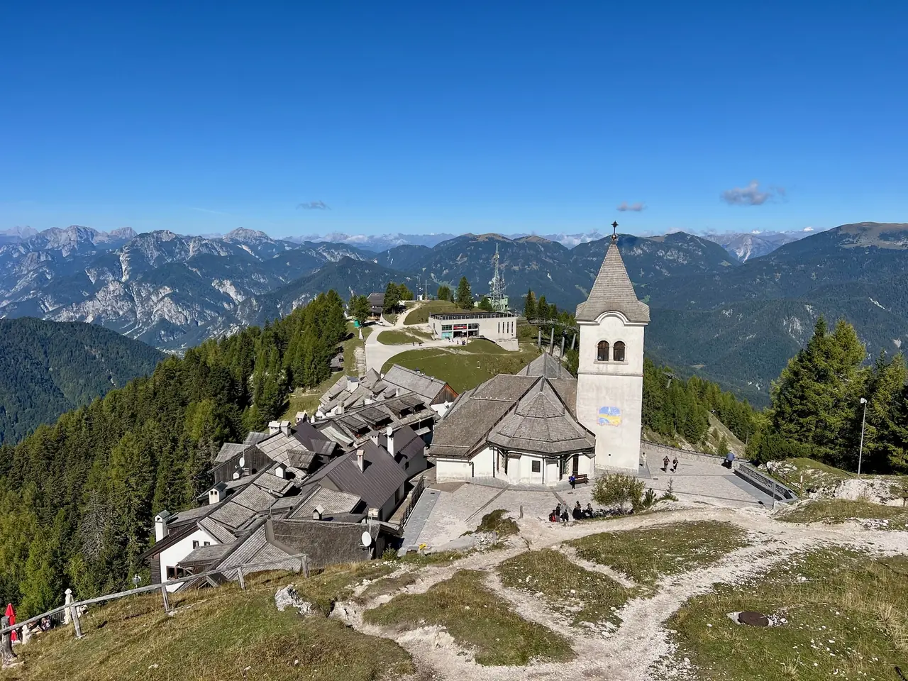 Pohled shora na šedé střechy osady Monte Lussari a zvonici svatyně, za nimi pásmo Julských Alp