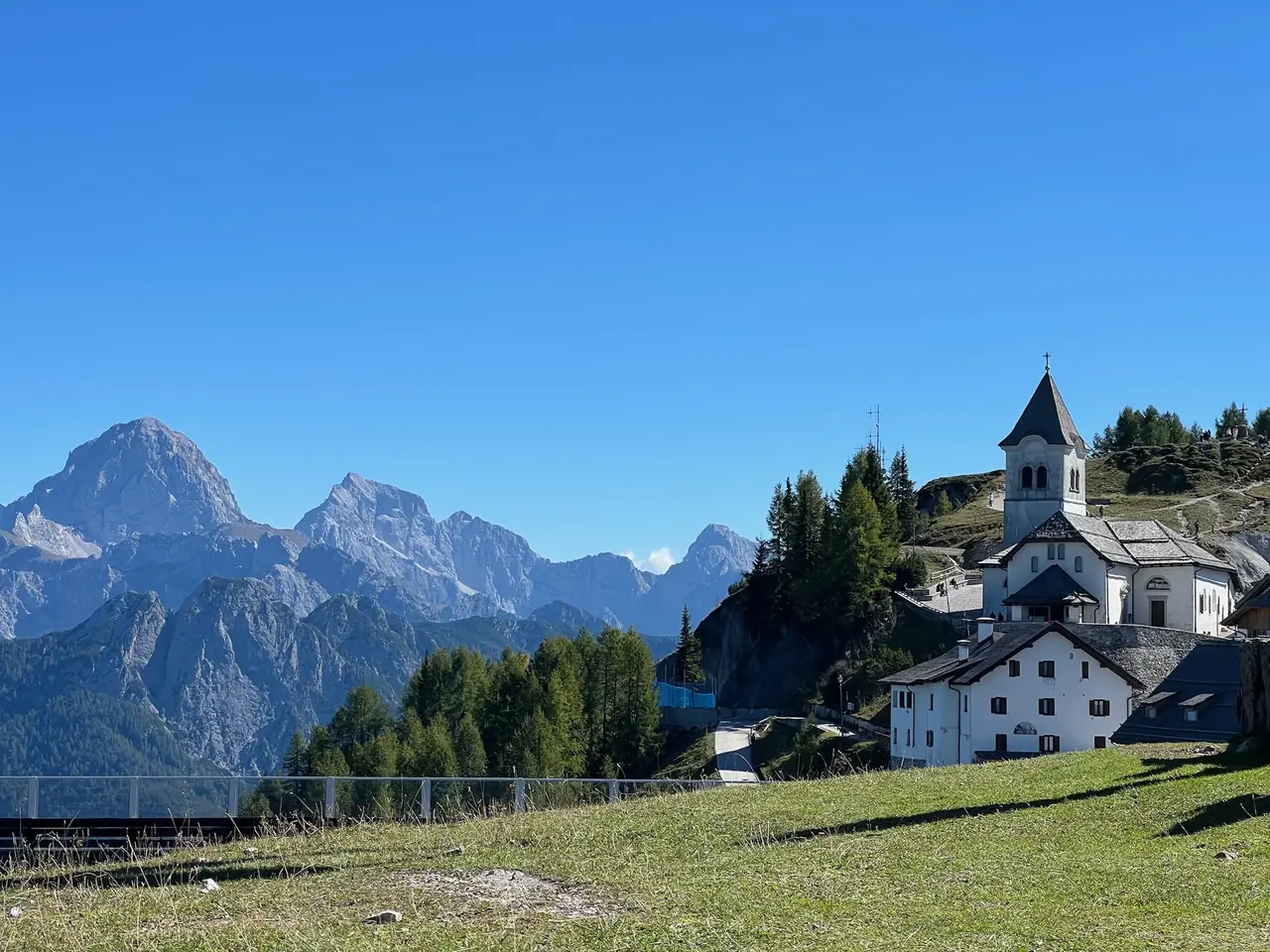 Horská vesnička Monte Lussari s bílou svatyní a zvonicí na zelené louce, v pozadí skalnaté štíty Julských Alp