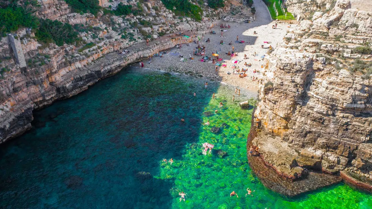 Letecký pohled na zátoku Polignano a Mare s průzračnou tyrkysovou vodou, kamennou pláží a bílými vápencovými útesy plnými koupajících se lidí