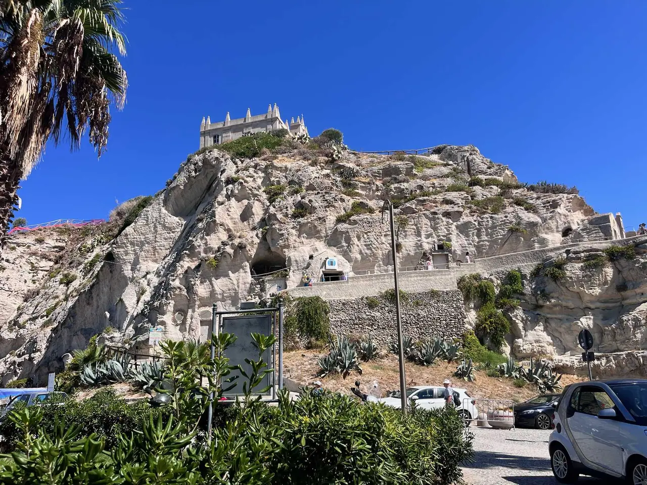 Bílý kostel Santa Maria dell'Isola na skále v Tropei s palmami a modrou oblohou