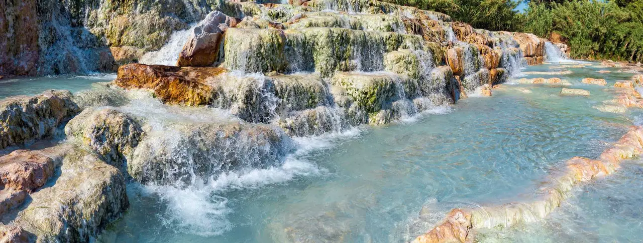Naše oblíbené termální bazénky v Saturnia s tyrkysovou vodou tekoucí přes bílé vápencové terasy mezi zelenými stromy