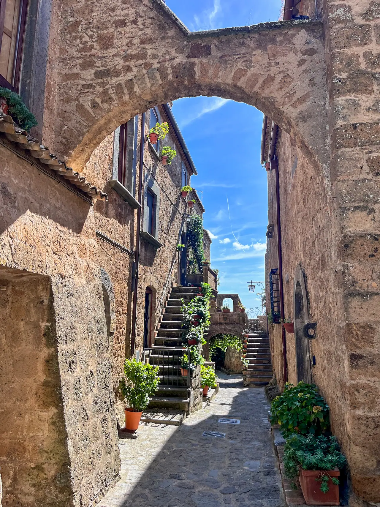Úzká kamenná ulička v Civita di Bagnoregio s květinami, schodišti a historickými domy.
