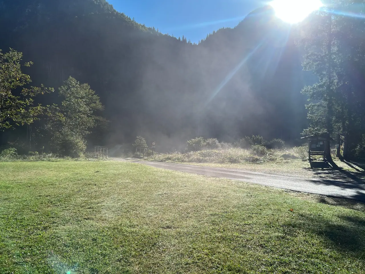 Ranní mlha stoupá nad loukou u parkoviště ve Via Saisera, za ní zalesněný kopec ve slunci