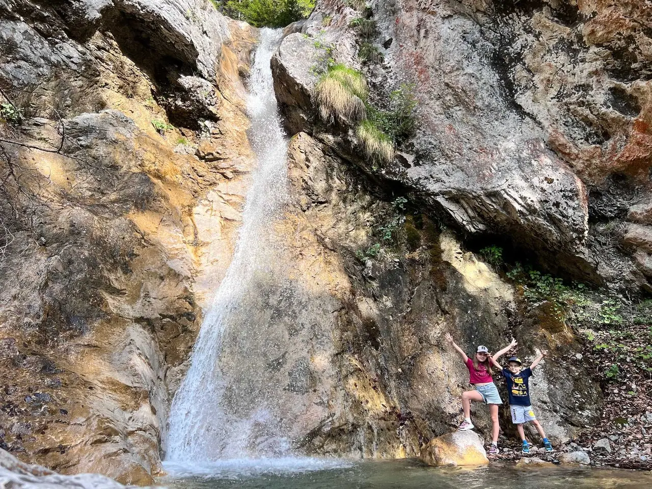 Naše děti pózují s rukama nahoře na skále před vodopádem v údolí Val Saisera