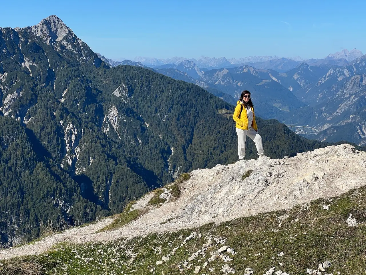Monika ve žluté bundě na skalní vyhlídce nad Monte Lussari se štíty Julských Alp v pozadí