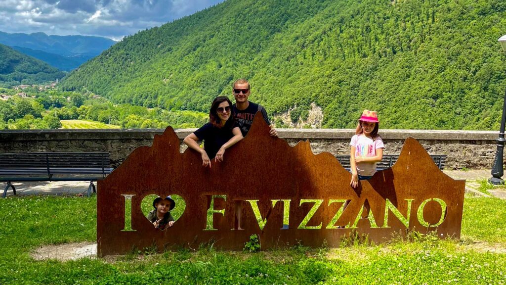 Rodina se fotí u velkého kovového nápisu I LOVE FIVIZZANO na vyhlídce s hornatou zelenou krajinou Toskánska v pozadí