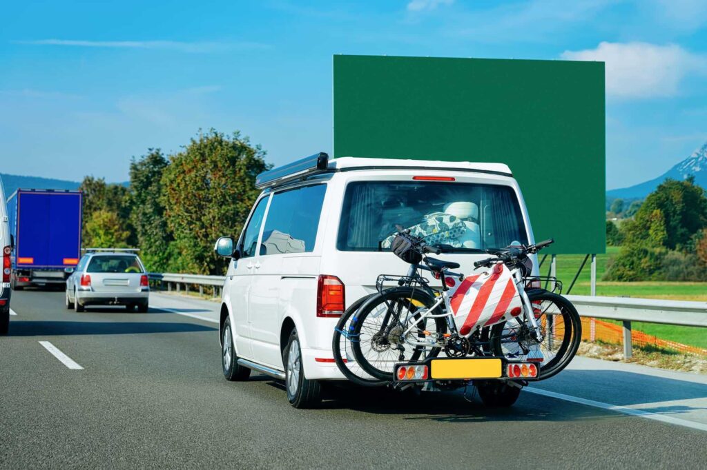 Bílé auto s nosiči kol na zadní části jede po dálnici, na kole je červeno-bílá výstražná tabule s reflexními pruhy