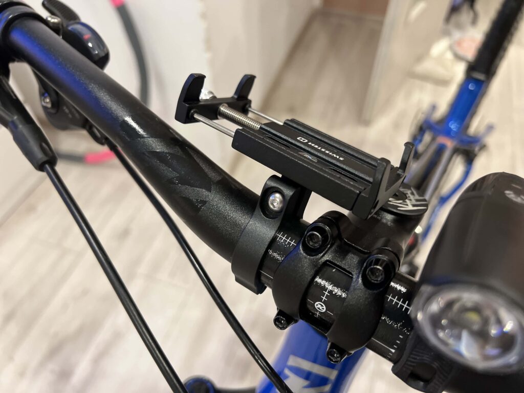 Detail černého kovového držáku telefonu Swissten BC2 připevněného na modrém rámu kola s nastavitelným mechanismem a šroubovacím upínáním