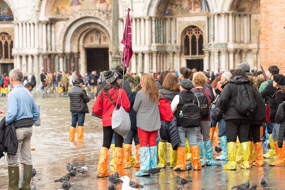 Turisté v barevných plastových návlecích na nohou brodí zaplavené náměstí sv. Marka před bazilikou během acqua alta v Benátkách