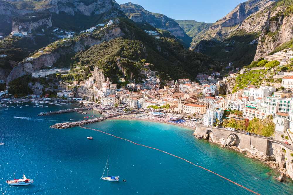 Pohled z moře na Amalfi s bílými domy tísněnými mezi skálami a tyrkysovou zátokou s jachetami