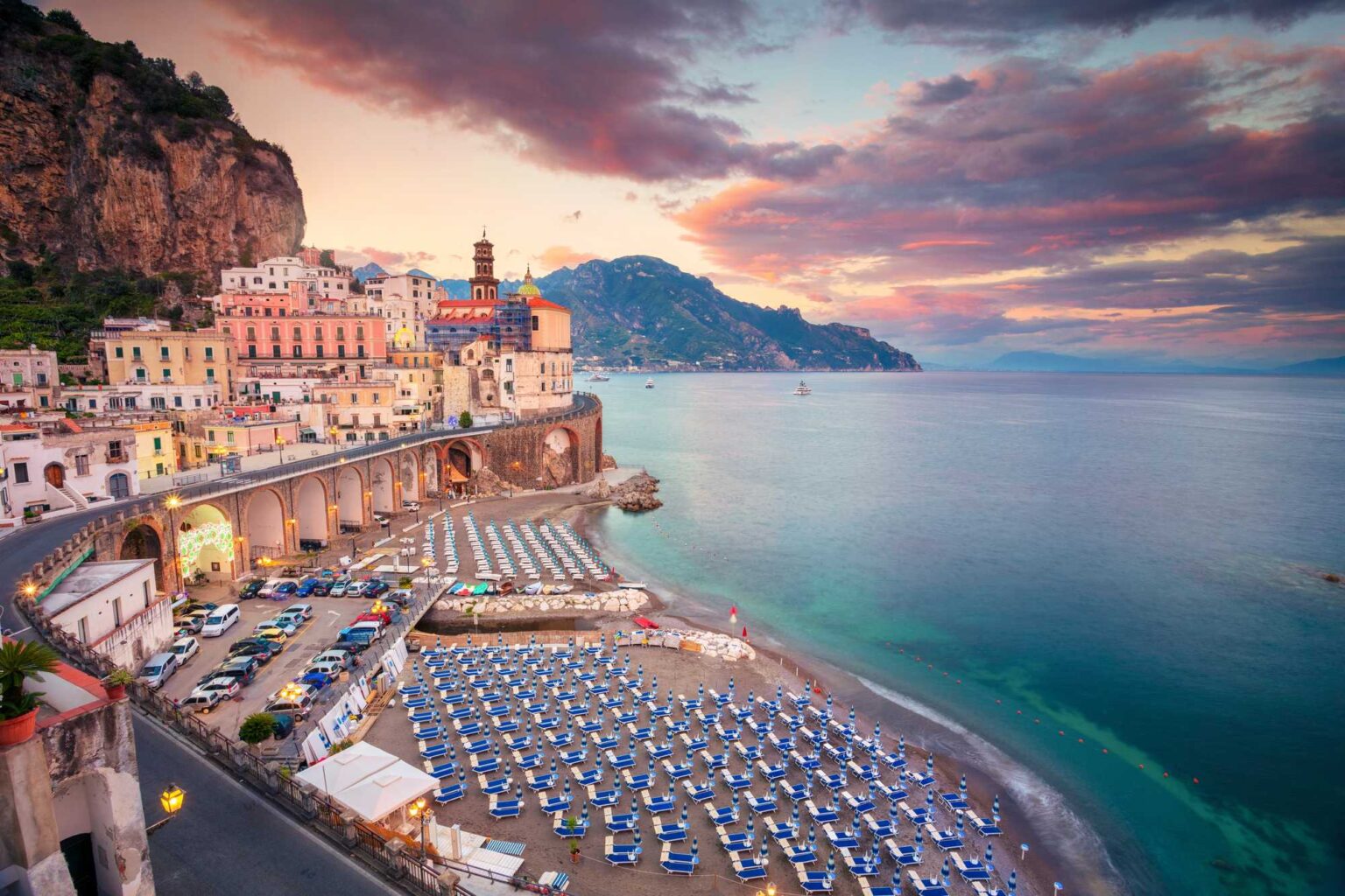 Pohled shora na Atrani na pobřeží Amalfi, kde řady modrých lehátek lemují tyrkysové moře pod růžovým večerním nebem a kamennými arkádami silnice