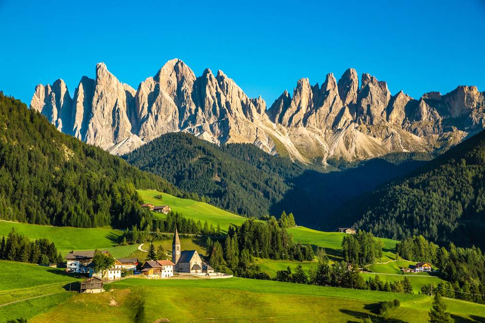Kostelík Santa Maddalena ve Val di Funes obklopený sytě zelenými loukami s jehlicovitými štíty Dolomit v pozadí, červenec bez davů.
