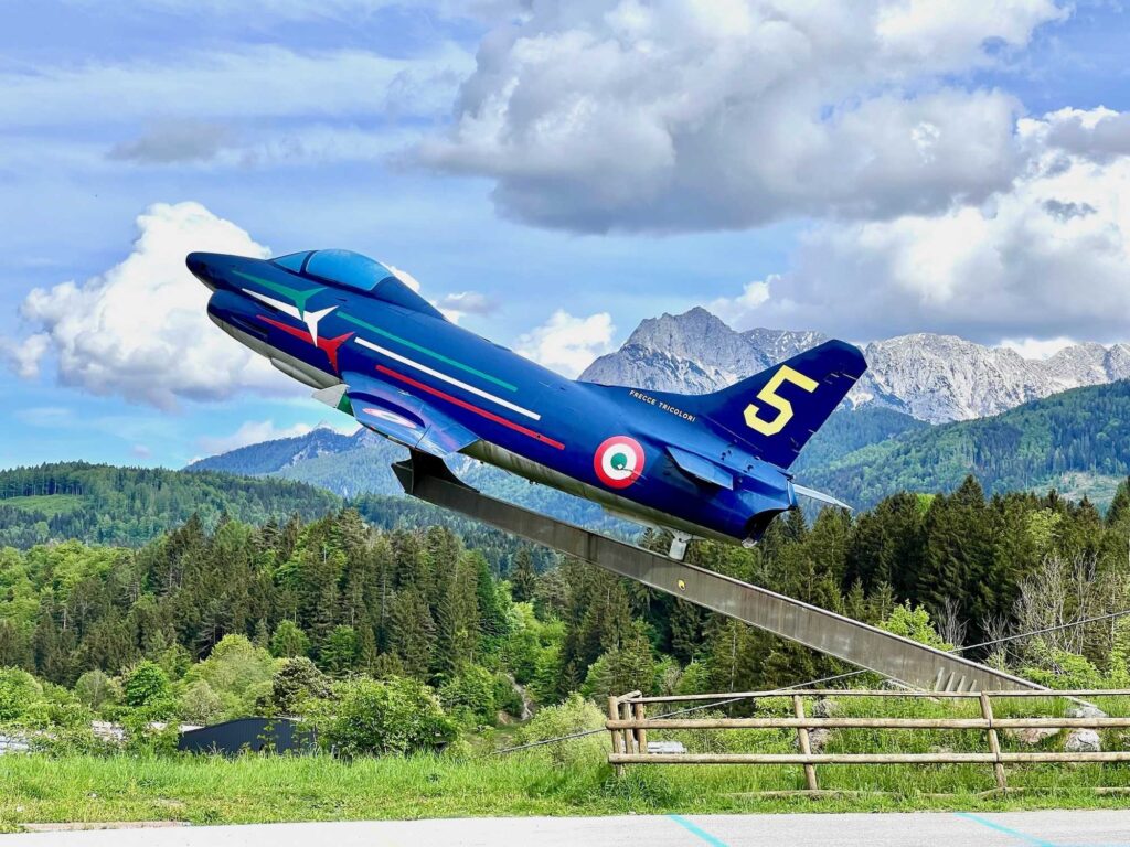 Modrý akrobatický letoun Frecce Tricolori číslo 5 na podstavci v údolí Val Saisera s Julskými Alpami v pozadí.