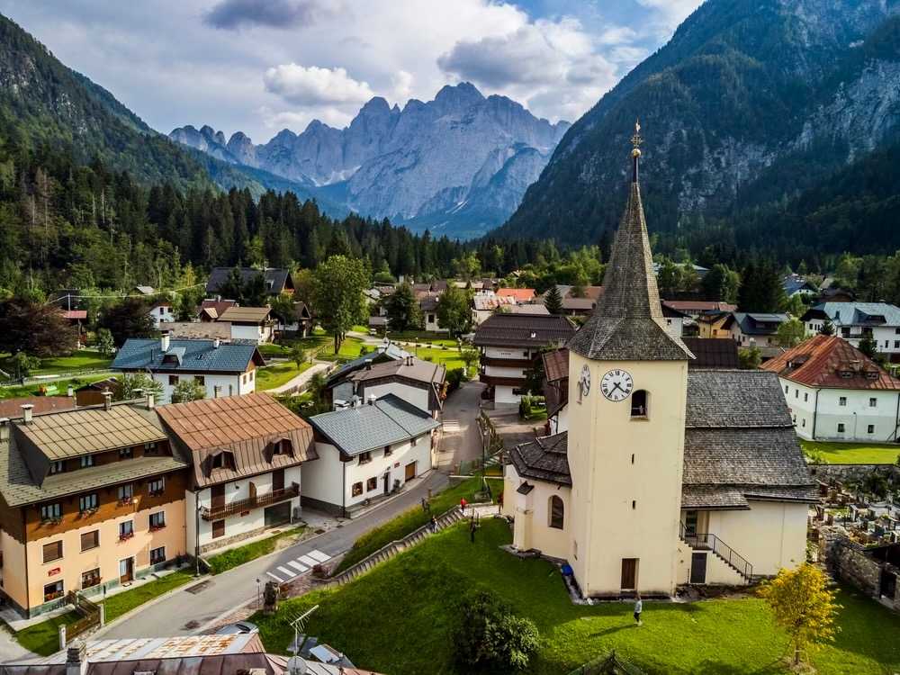 Letecký pohled na vesnici Malborghetto-Valbruna s bílou věží kostela s hodinami, obklopenou alpskými domky a skalnatými vrcholy údolí Val Saisera