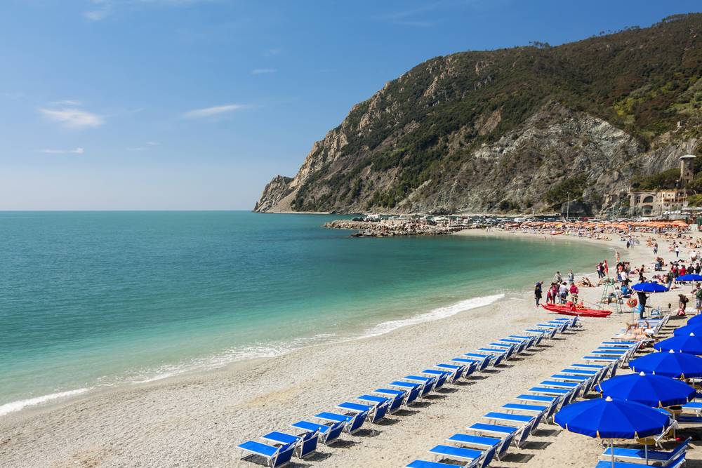 Pláž Monterosso al Mare s řadami modrých lehátek a slunečníků, tyrkysové moře a skalnatý kopec v pozadí