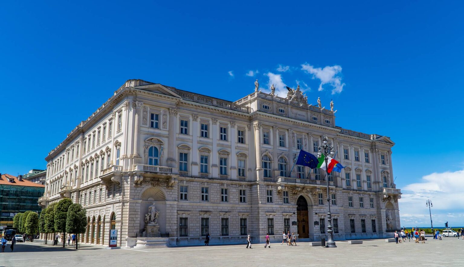 Bílý barokní palác na náměstí Piazza Unità d'Italia v Terstu s vlajkami EU a Itálie za jasného letního dne