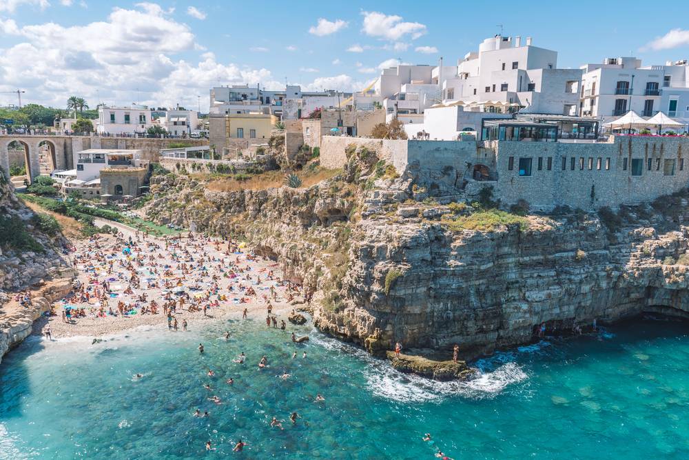 Přeplněná pláž v Polignano a Mare s tyrkysovým mořem, bílými domy na vápencových útesech a koupajícími se lidmi