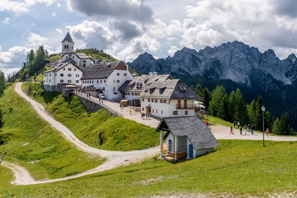 Bílý kostel a alpské ubytovny na zeleném vrcholu Monte Lussari nad údolím Val Saisera s kamennou cestou vedoucí k budovám