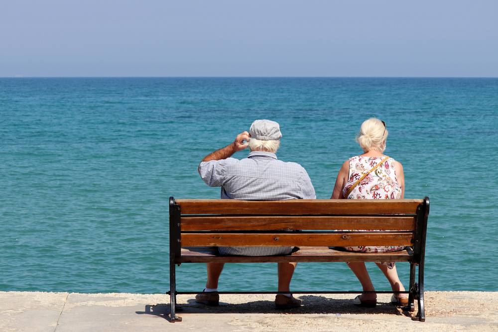 Starší manželský pár sedí zády na dřevěné lavičce u italského tyrkysového moře a pozoruje klidný horizont za slunečného dne.