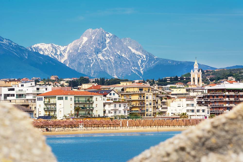 Italské přímořské město Alba Adriatica s plážovými slunečníky z palmového listí, bílé budovy s balkóny a zasněžený Gran Sasso v pozadí