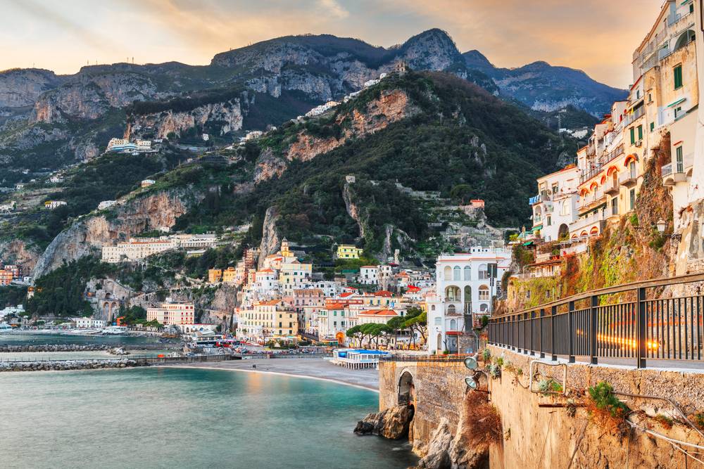 Amalfi při západu slunce – bílé domy přilepené na skalnatých útesech nad tyrkysovým mořem, cestička s kovým zábradlím vede podél skály