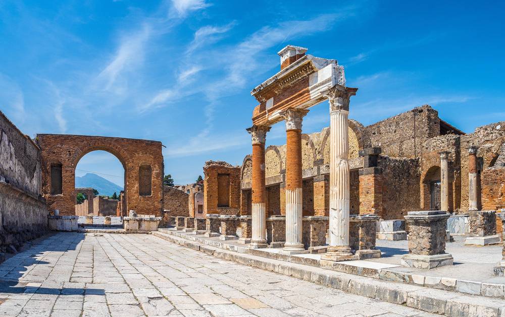 Antické sloupy a cihlové oblouky na hlavním náměstí Pompejí s horou Vesuv v pozadí za slunečného dne
