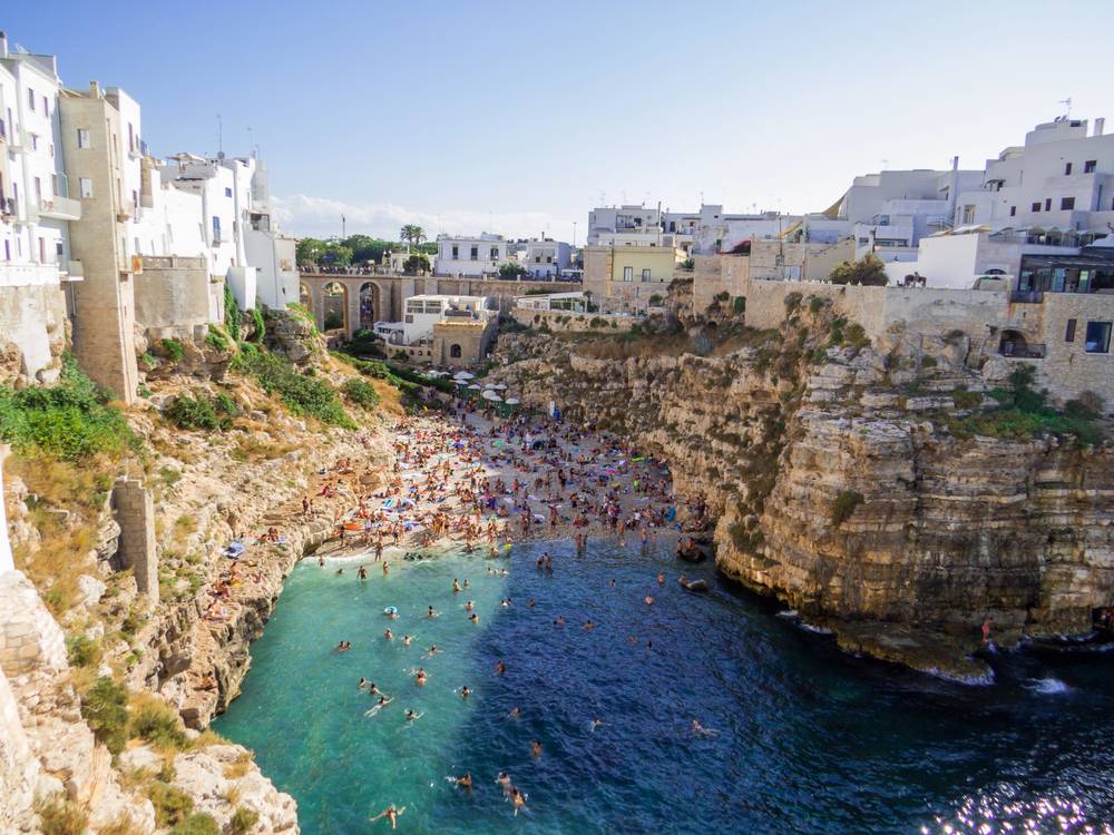 Přeplněná pláž Lama Monachile v Polignano a Mare sevřená vápencovými útesy s bílými domy na vrcholu skal