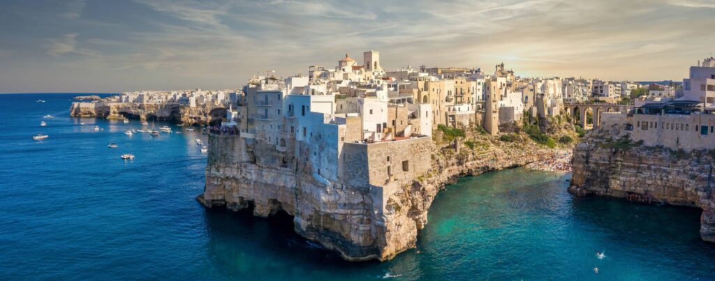 Bílé a kamenné domy starého města Polignano a Mare postavené přímo na vápencových útesech nad tyrkysovým Jaderským mořem, lodičky plují kolem skal