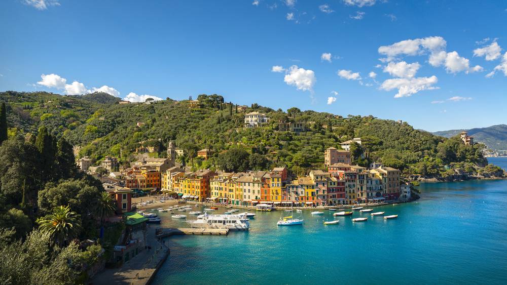 Portofino přístav s jachtami na tyrkysové vodě, barevné italské domy oranžové a žluté podél zálivu, zelené zalesněné kopce v pozadí
