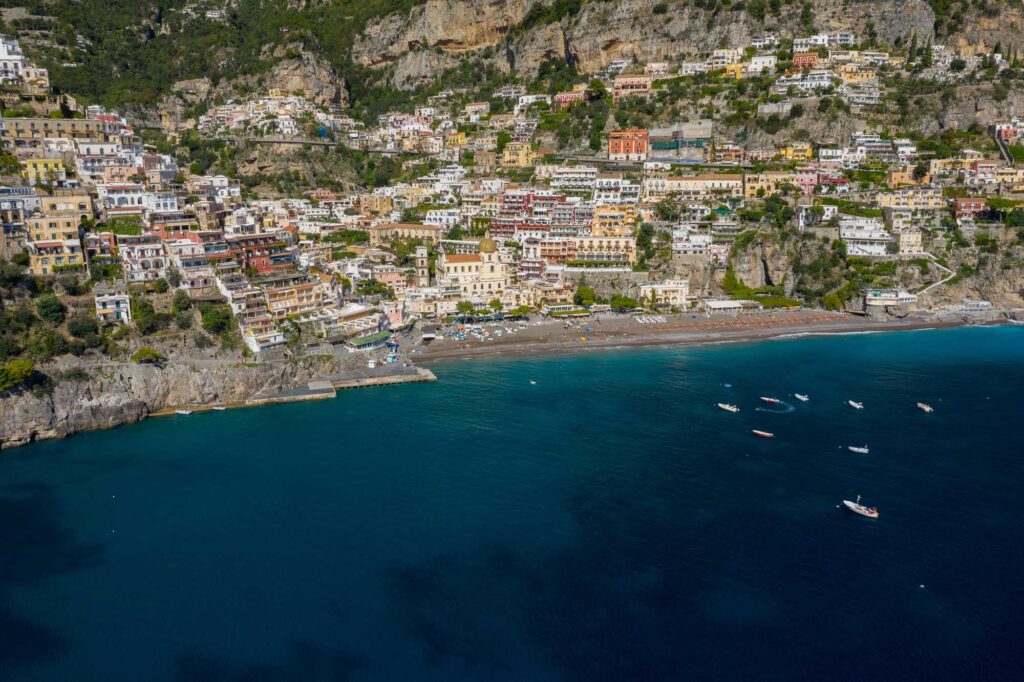 Positano z ptačí perspektivy – barevné domy naskládané na skalnatém útesu nad temně modrou hladinou Tyrhénského moře s kotvícími loďkami