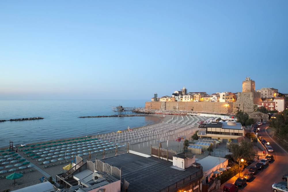 Termoli při soumraku - pláž Sant Antonio s řadami zavřených slunečníků, středověká pevnost a přístav s tradičním traboccem v pozadí