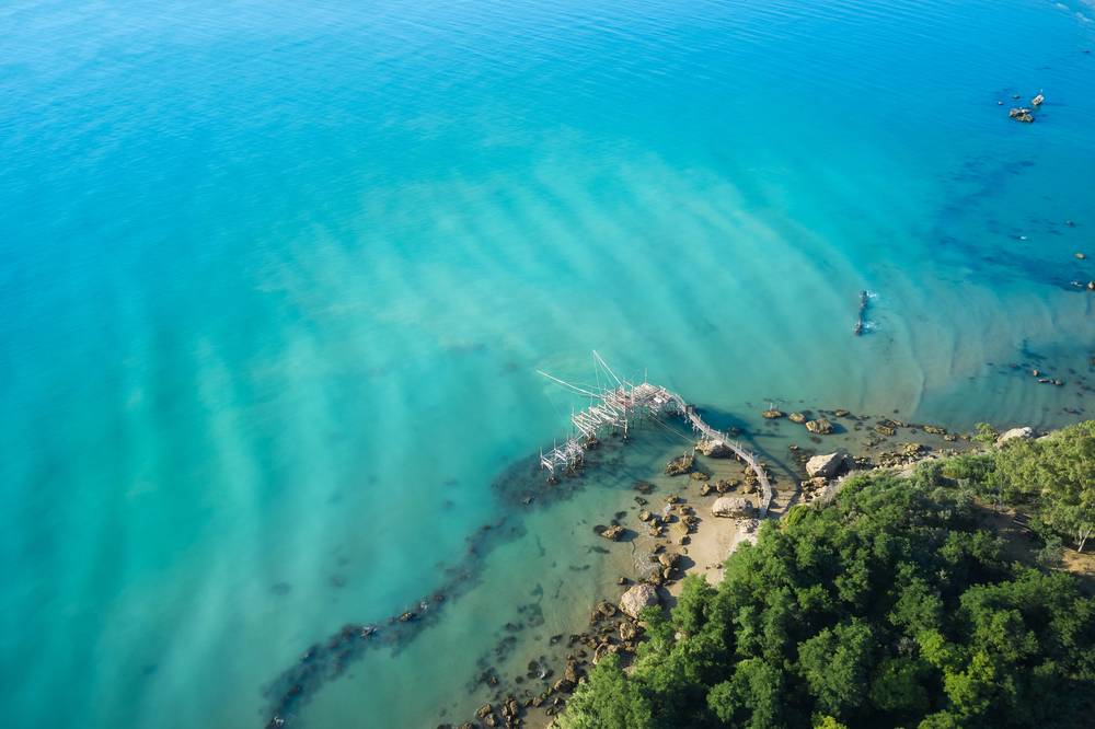Letecký pohled na tradiční rybářské molo trabocco u pobřeží Marina di Vasto v Abruzzu, obklopené tyrkysovým Jaderským mořem a skalnatým břehem porostlým zelení.