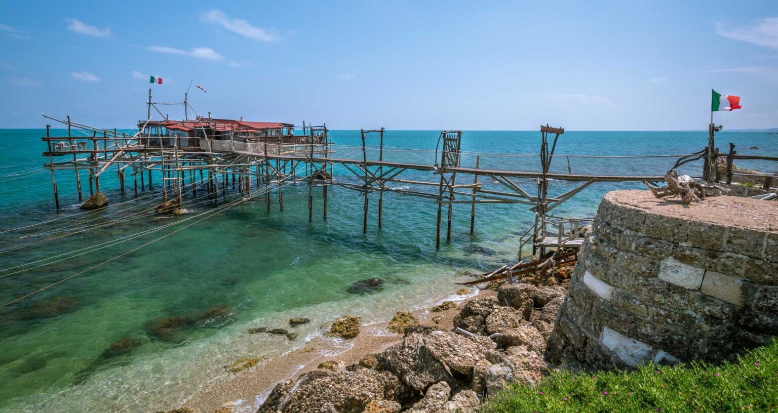 Tradiční dřevěná rybářská platforma trabocco na Jadranském moři s italskými vlajkami a tyrkysovou vodou v Abruzzách