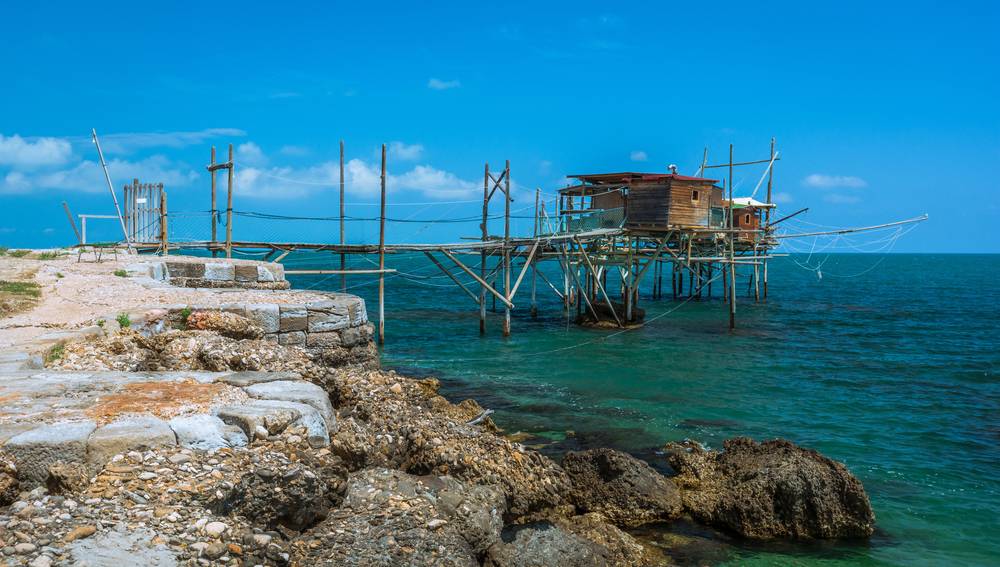 Tradiční dřevěná rybářská věž trabocco na kůlech nad tyrkysovým Jaderským mořem, pobřeží Abruzzo s kamennými útesy v popředí