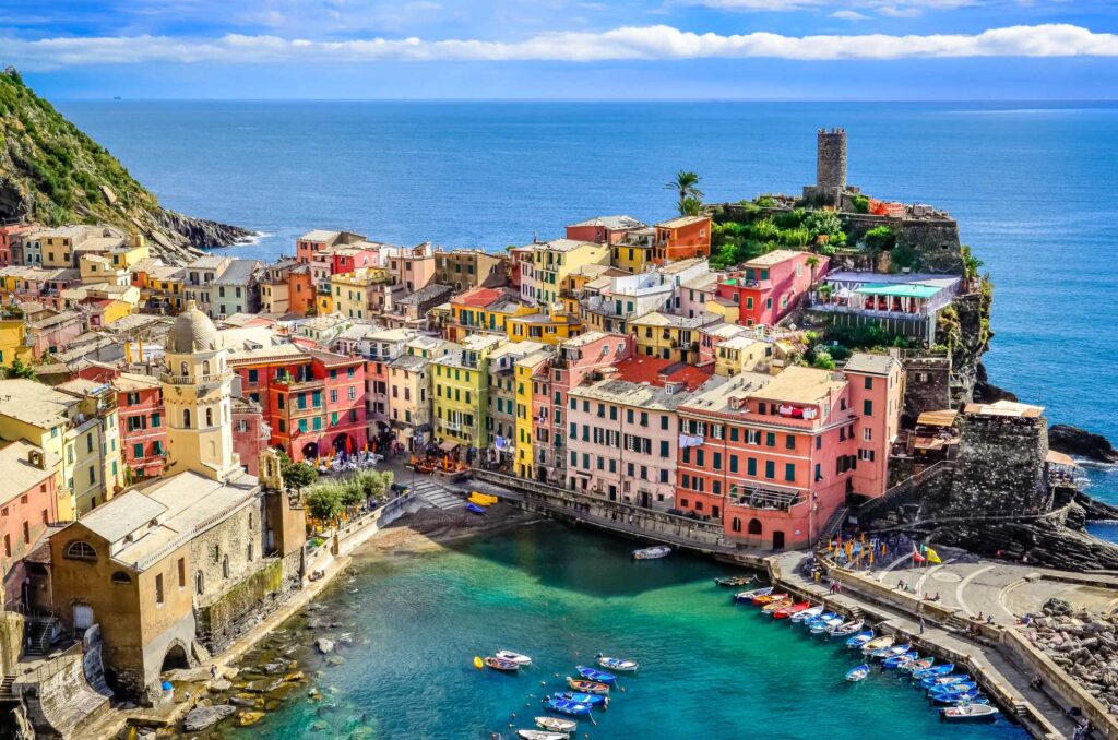 Vernazza v Cinque Terre shora – pestrobarevné domy narůžovělých a žlutých fasád tísněné nad tyrkysovým přístavem s loďkami